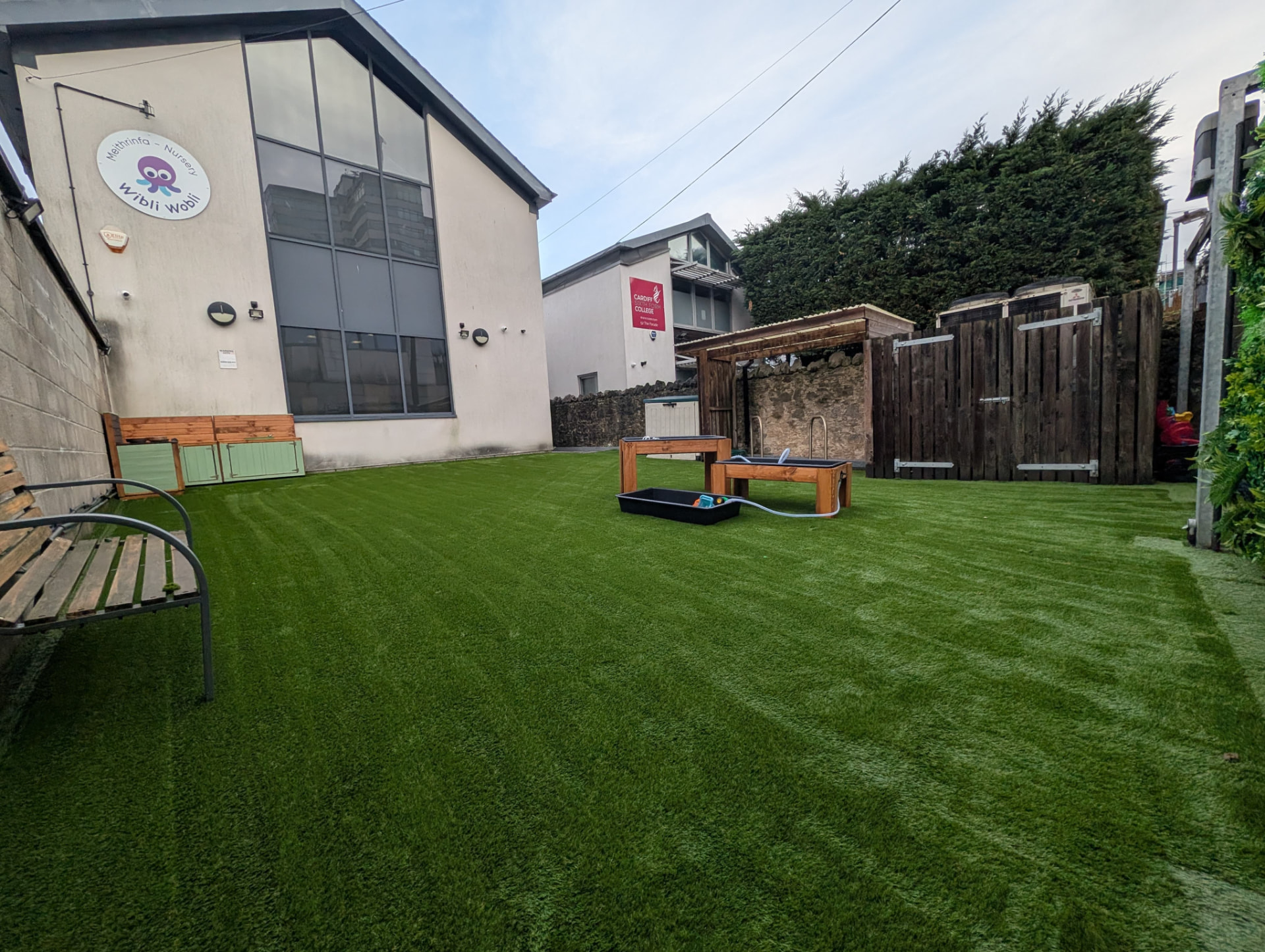 Secure nursery garden at Wibli Wobli Cardiff with artificial grass, outdoor play equipment and the nursery building
